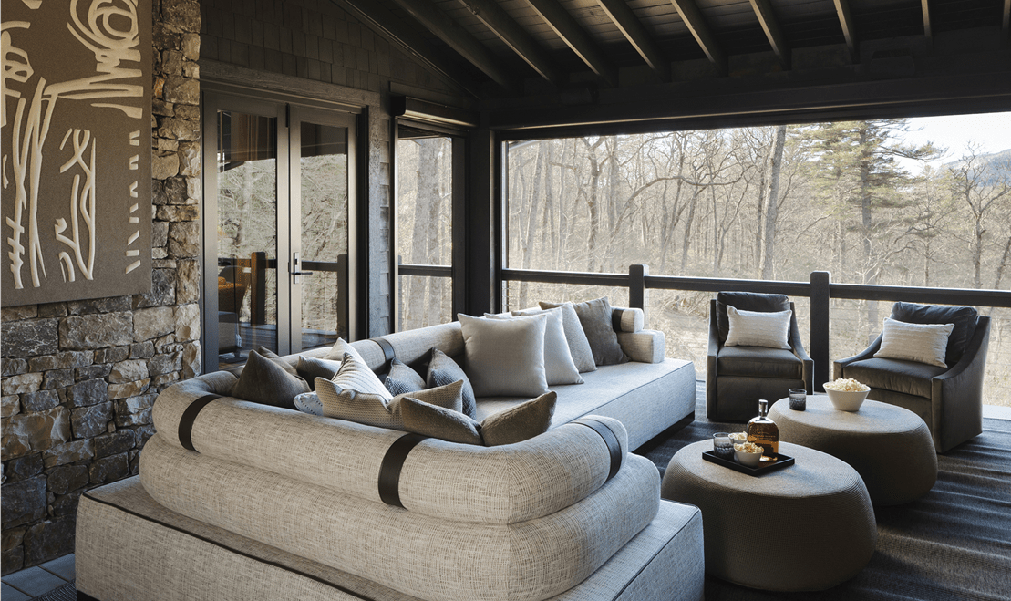 An outdoor patio with a sectional sofa, two armchairs, and two ottomans on a covered deck overlooking a forest.
