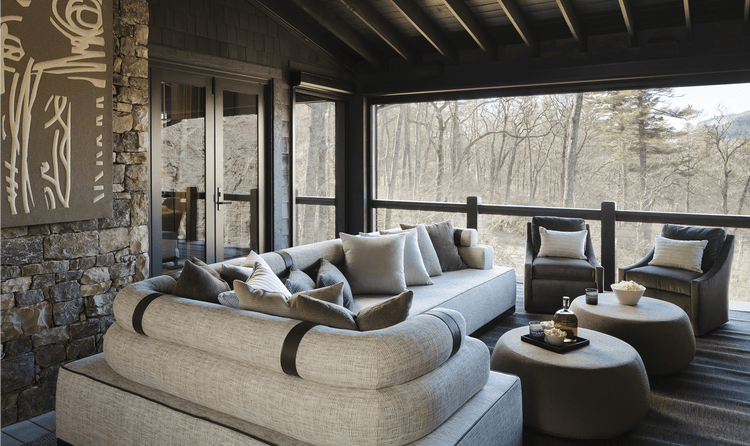 An outdoor patio with a sectional sofa, two armchairs, and two ottomans on a covered deck overlooking a forest.