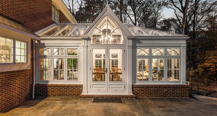 A beautiful, white all-season room with large windows covering the ceiling and walls connected to a brick, luxury home.