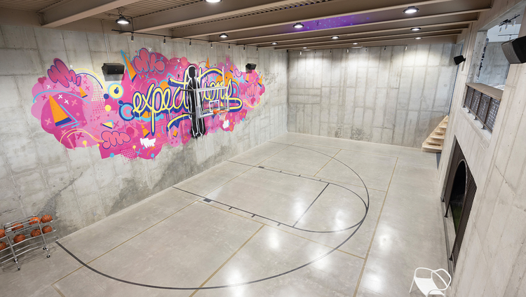 Beneath the garage sits this basketball half court with grey walls and colorful graffiti behind the main hoop.