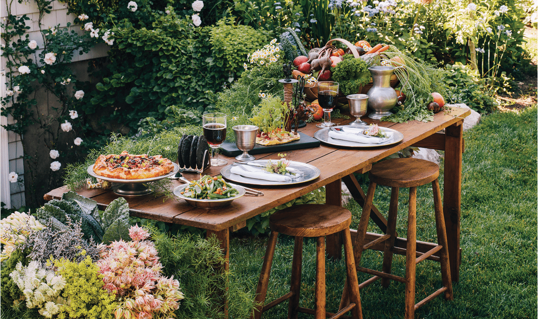 An antique table set outside with wooden chairs and meals spread on the table-top with a garden behind it.