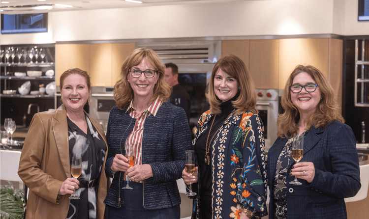 Four individuals posed in front of Sub-Zero, Wolf, and Cove appliances with a drink in their hands at the Riggs showroom.