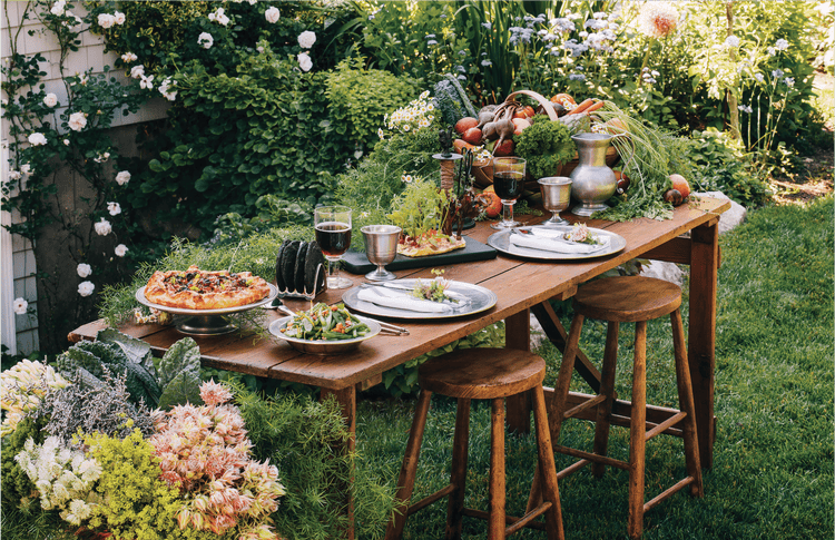 An antique table set outside with wooden chairs and meals spread on the table-top with a garden behind it.