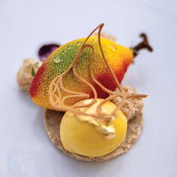 A fruit-shaped mousse with red, green, and yellow hues on a biscuit base, topped with sugar art and gold leaf.