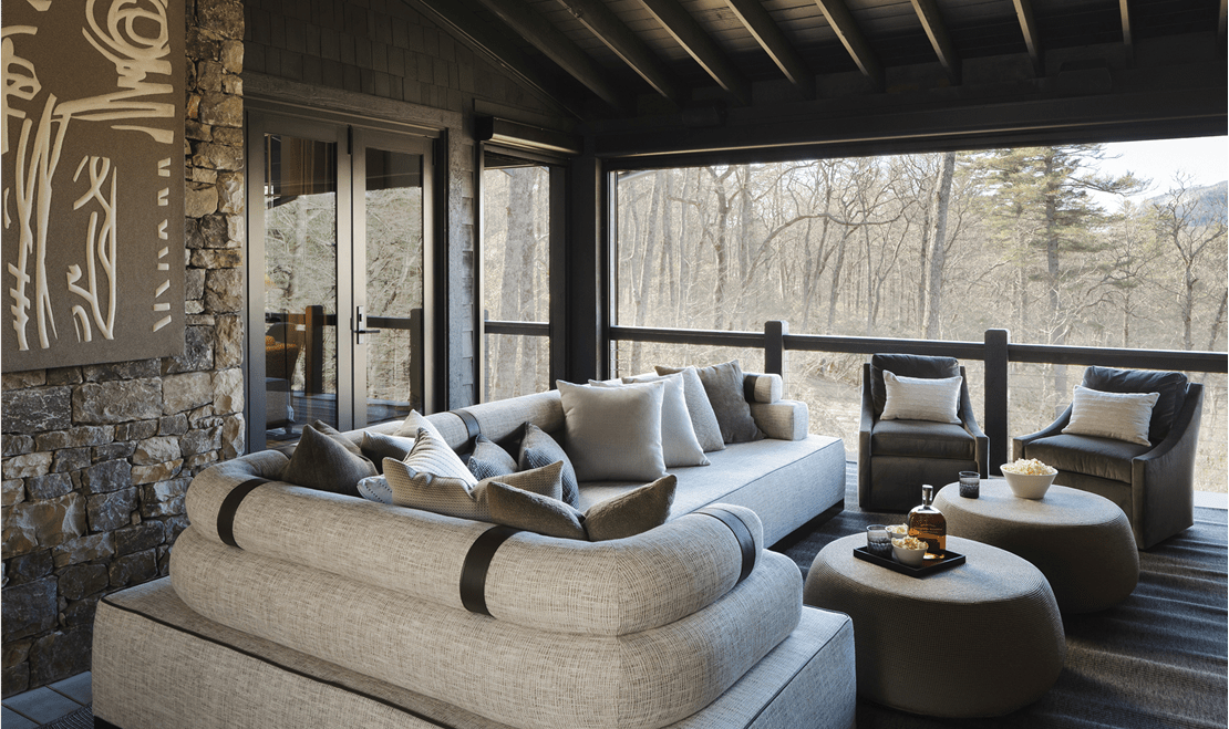 An outdoor patio with a sectional sofa, two armchairs, and two ottomans on a covered deck overlooking a forest.