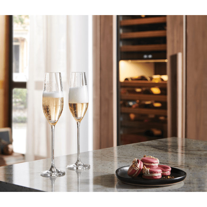 Two champagne glasses sitting on a marble table in front of a luxury wine refrigerator.