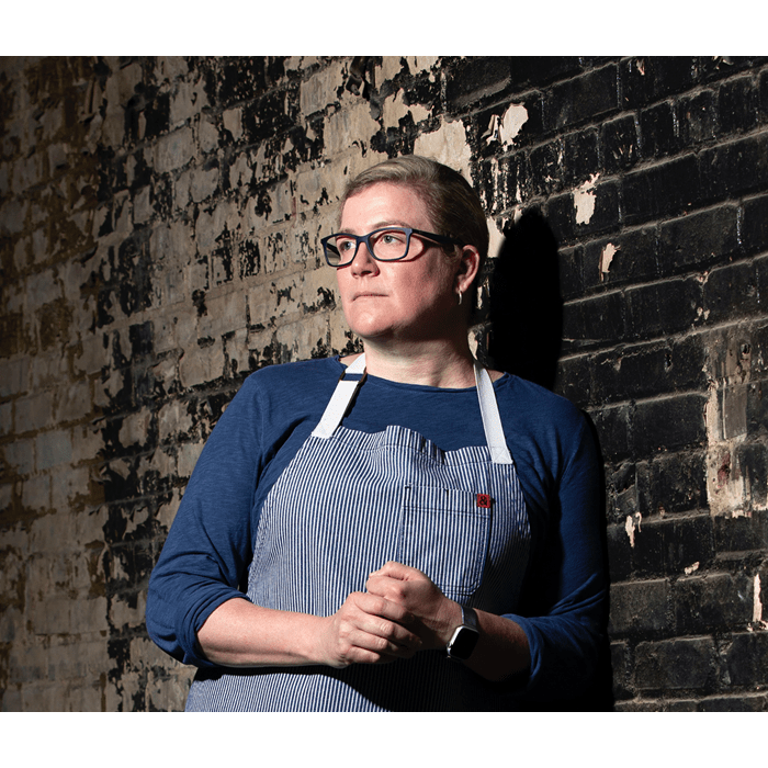 Chef Ashley Christensen poses in front of a brick wall in an apron with her hands clasped together.