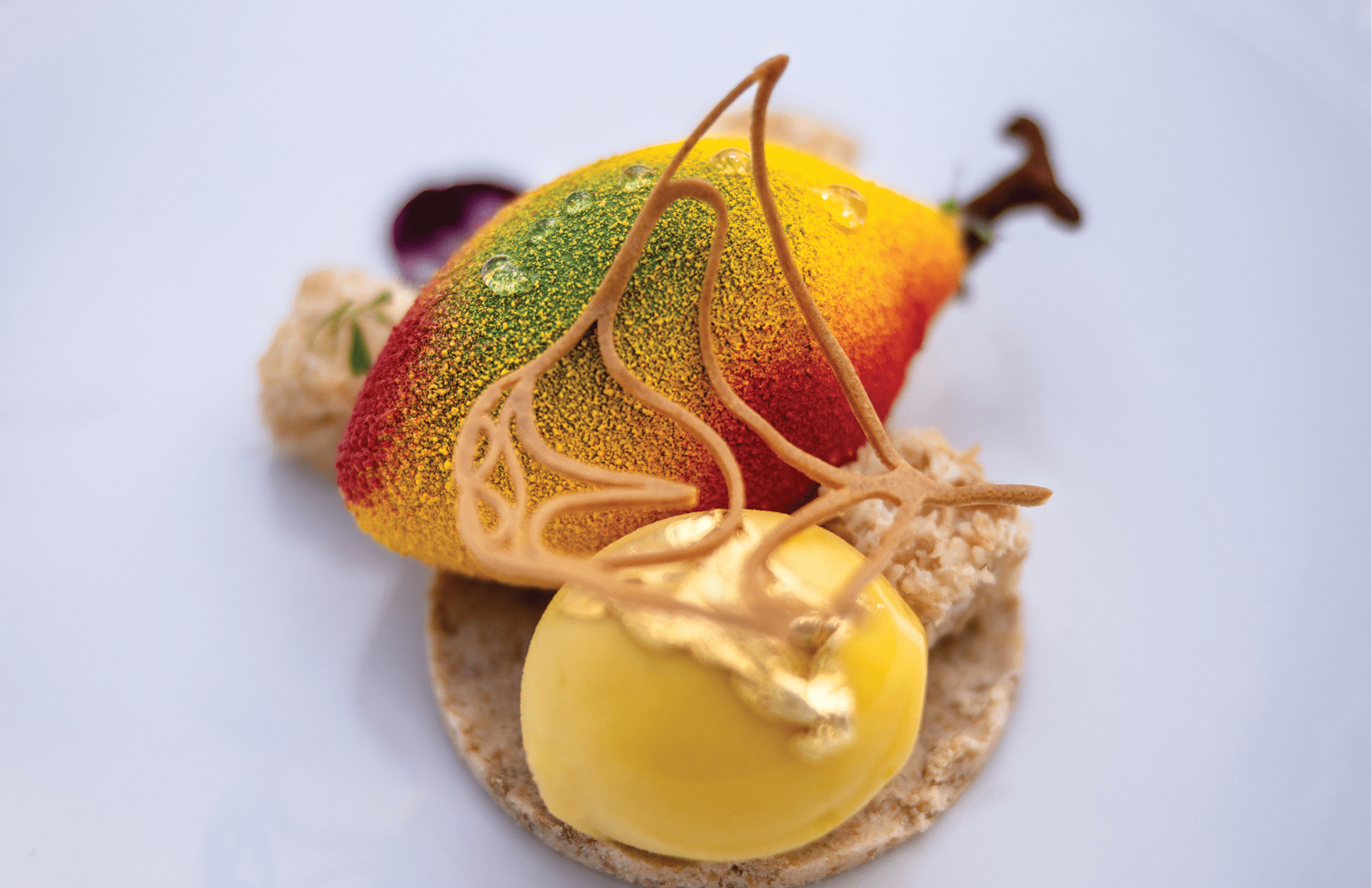 A fruit-shaped mousse with red, green, and yellow hues on a biscuit base, topped with sugar art and gold leaf.