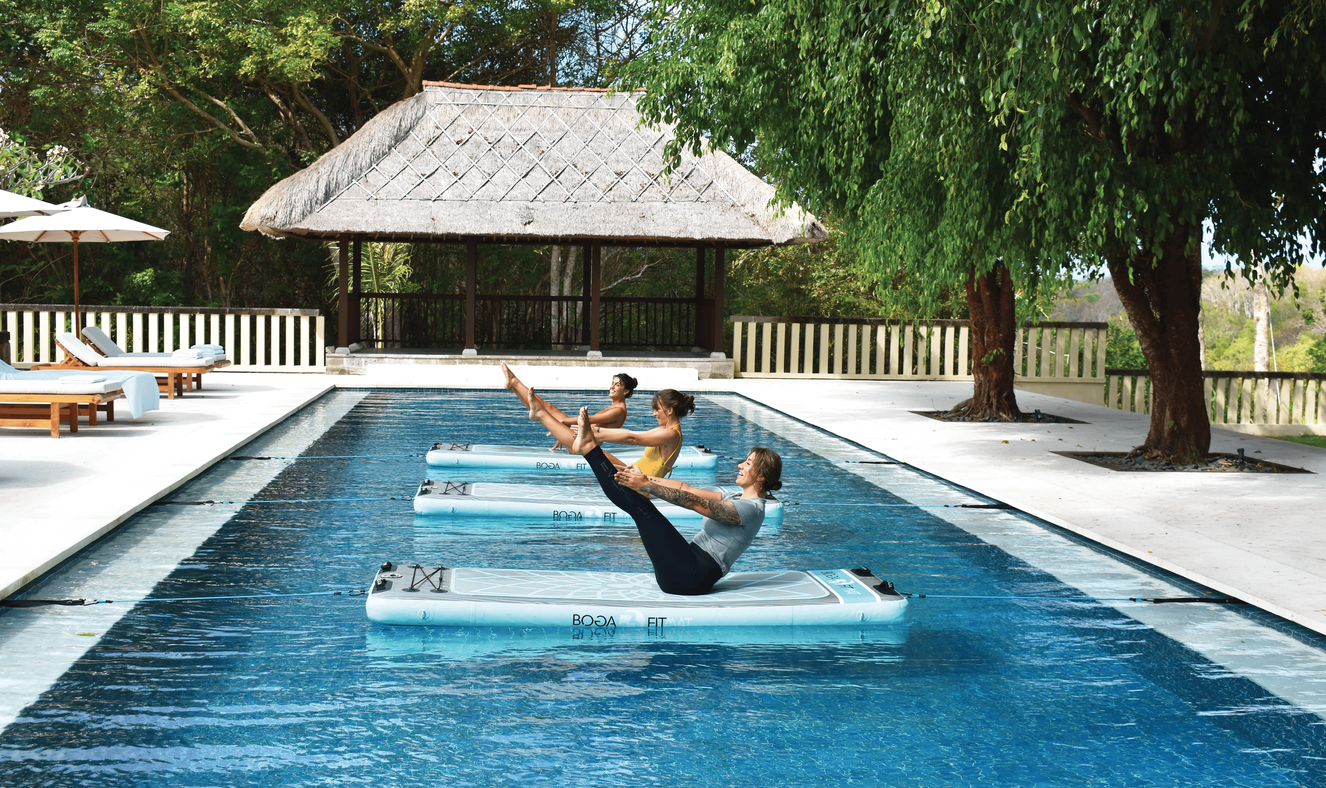 Three individuals doing yoga on boards in an outdoor pool at the REVĪVŌ Wellness Resort in Bali, Indonesia.