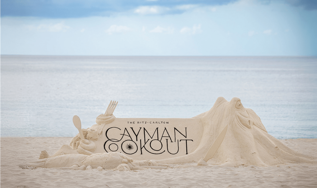 A sand sculpture with "The Ritz-Carlton Cayman Cookout" written on top of it in black.