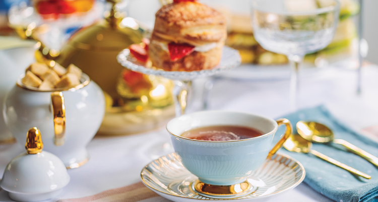 A small cup of tea in front of a breakfast dessert and sugar cubes on a white tablecloth.
